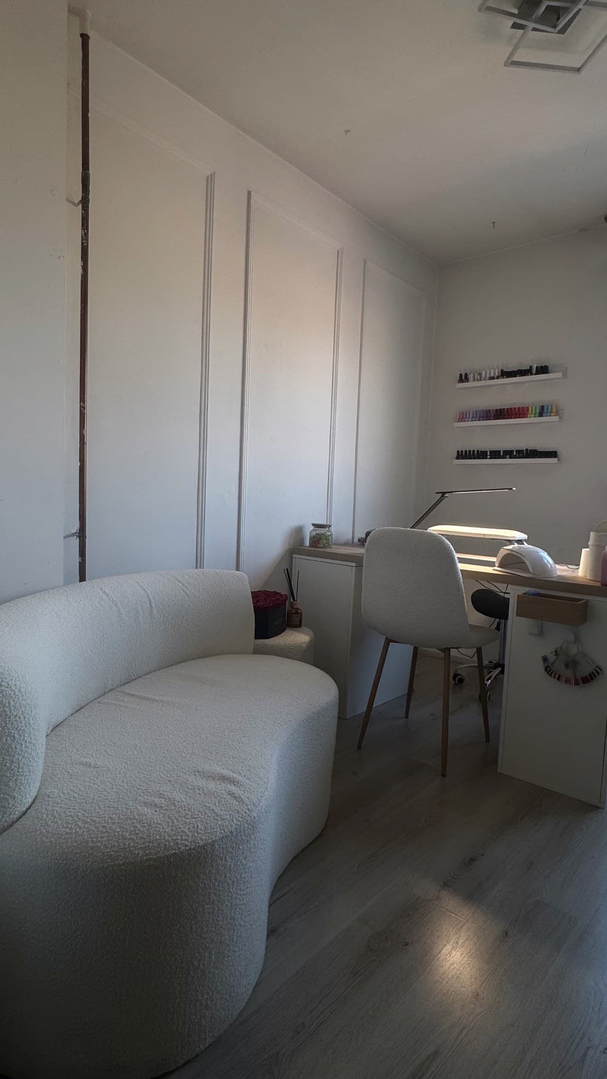 De salon van The Nail Bar by Eve in Tegelen: witte bouclé-bank, werkplek met lamp en een wand vol nagellak